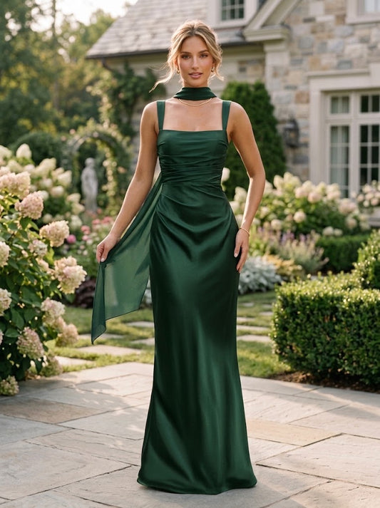 A woman stands outdoors in a formal garden wearing a floor-length dark green satin gown with a high neck, square straps, ruched bodice, and a draped side panel. The background shows stone house architecture, greenery, and flowering plants.