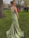 Woman in a green evening gown with a open back standing on grass with trees and people in the background.